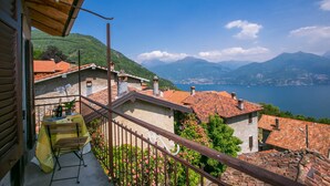 A balcony with a table, chairs, and a bottle of wine. The balcony overlooks a scenic view of a lake and mountains. - Plesio Rustico Panoramica (Plesio)
