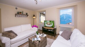 A living room with a white sofa, a wicker coffee table with a book, a TV on a stand, and a window with a mountain view. - Plesio Rustico Panoramica (Plesio)