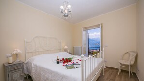 A bedroom with a bed, bedside tables, a chair, a chandelier, and a view of the outdoors. - Plesio Rustico Panoramica (Plesio)