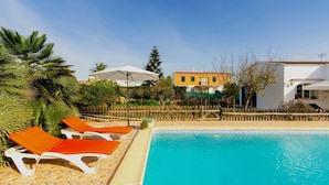 Une piscine entourée d’une clôture en bois et de verdure, avec des chaises longues et des parasols.
