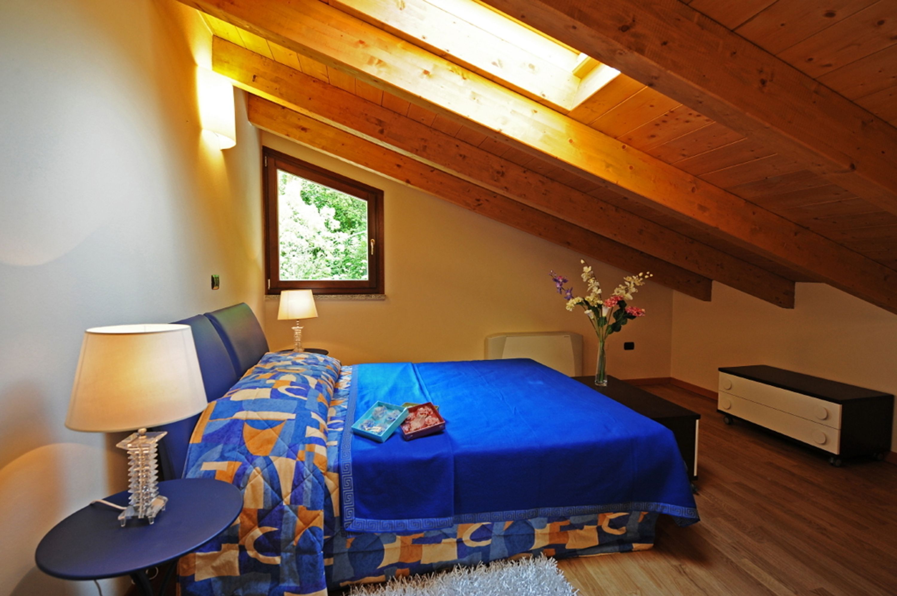A bedroom with a sloped ceiling, a bed with a blue bedspread, a bedside table with a lamp, a small table, and a nightstand with a vase of flowers.