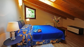 Una camera da letto con soffitto spiovente, un letto con copripiumino blu, un comodino con una lampada, un piccolo tavolino e un altro comodino con un vaso di fiori.