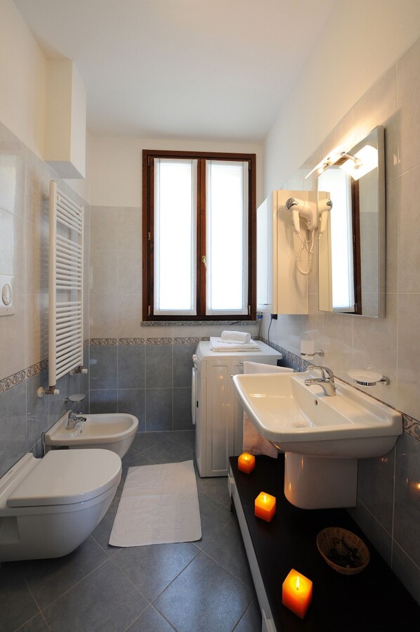 A modern bathroom with a toilet, bidet, and sink. There is a window above the bidet, a towel rack, and a mirror with a telephone. - Residence Eden Magnolia 16 (Argegno)