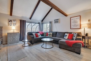 Un salon moderne avec un canapé d’angle, deux petites tables rondes, une cheminée et de grandes fenêtres.