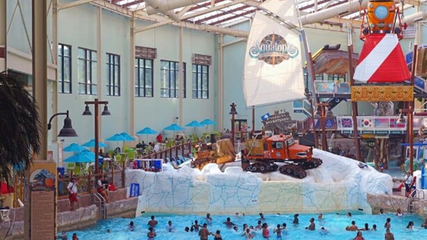 Ein großer, überdachter Wasserpark mit Schwimmbad, einem Spielbereich im Stil eines Schiffswracks und einem Rettungsschwimmerstand.