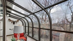 Ein Wintergarten mit einer gewölbten Glasveranda, ein rotes gepolstertes Sessel, ein kleiner Tisch mit einer Lampe sowie ein aufgehängter Tierkopf.