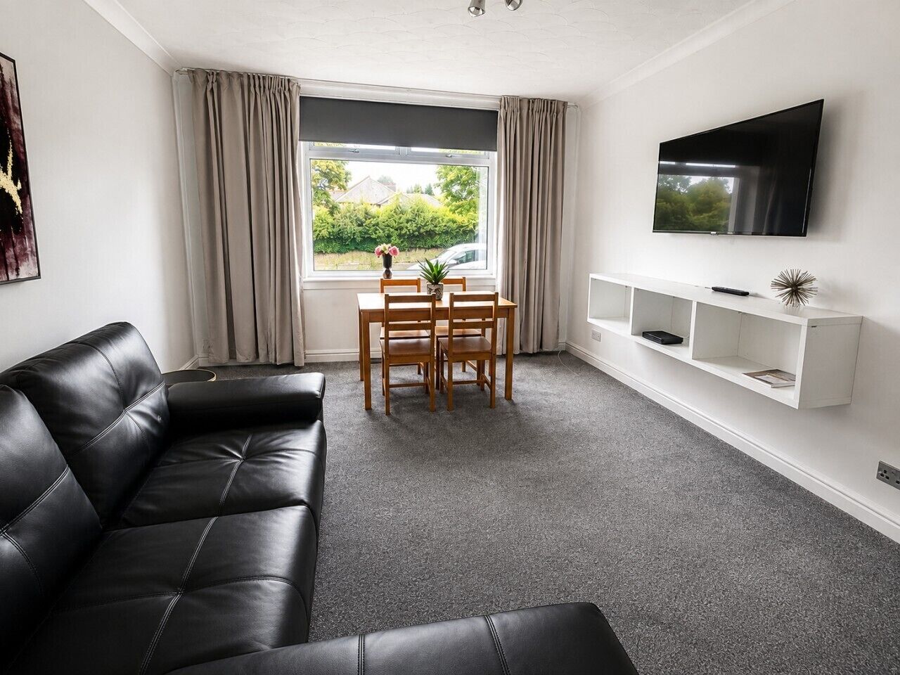 Een moderne woonkamer met een zwarte leren bank, een eettafel met stoelen, een aan de muur bevestigde televisie en een raam met gordijnen.