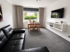 A modern living room with a black leather sofa, a dining table with chairs, a TV mounted on the wall, and a window with curtains.