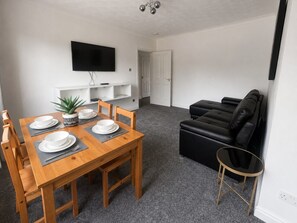 Ein Essbereich mit Holztisch, gedeckt für vier Personen, einer schwarzen Ledersofa und einem an der Wand befestigten Fernseher.