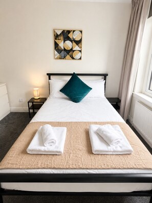 Ein Hotelzimmer mit Bett, Nachttisch, Lampe, einem Gemälde an der Wand und einem Fenster mit Vorhängen.