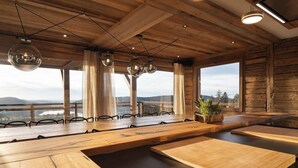 A wooden interior with a long table and chairs, large windows with curtains, and a view of the outdoors. - Alta Rocca, 14-guest chalet in Gérardmer – pool, jacuzzi, sauna (Gérardmer)