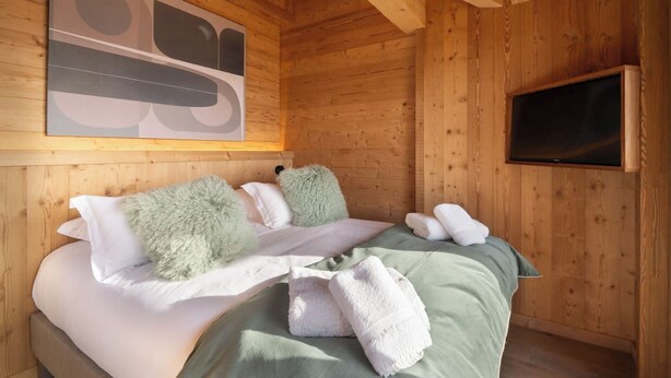 A wooden cabin room with a bed, pillows, a blanket, and a television. - Alta Rocca, 14-guest chalet in Gérardmer – pool, jacuzzi, sauna (Gérardmer)