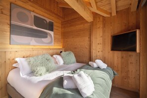 A wooden cabin room with a bed, pillows, a blanket, and a television. - Alta Rocca, 14-guest chalet in Gérardmer – pool, jacuzzi, sauna (Gérardmer)