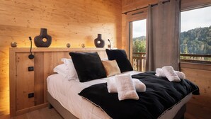 A wooden cabin bedroom with a bed, pillows, and a view of the forest. - Alta Rocca, 14-guest chalet in Gérardmer – pool, jacuzzi, sauna (Gérardmer)