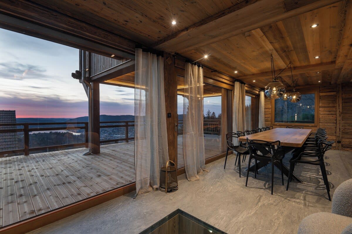 A spacious room with a large wooden table and chairs, large windows with curtains, and a view of the landscape.