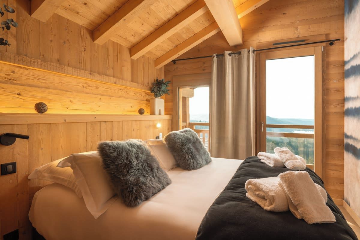 Une chambre dans un chalet en bois, avec un lit, des oreillers, des serviettes et une fenêtre donnant sur un paysage agréable.