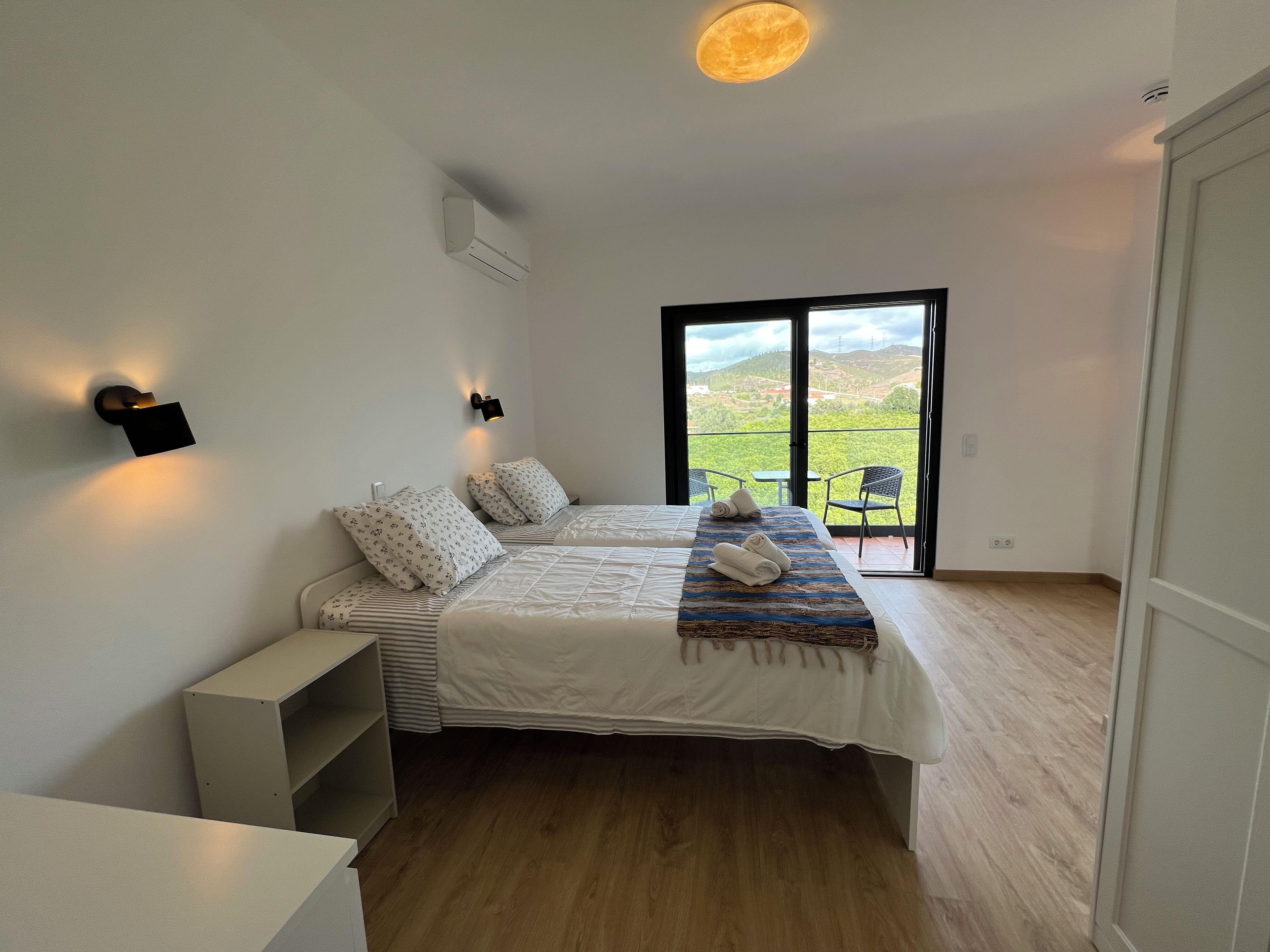 A bedroom with a bed, a nightstand, a desk, a chair, and a balcony with a view.