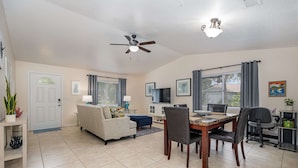 A spacious living room with a sofa, dining table, and chairs, a ceiling fan, and a TV. - Beachy Retreat 1 Mile to Beach (Satellite Beach)