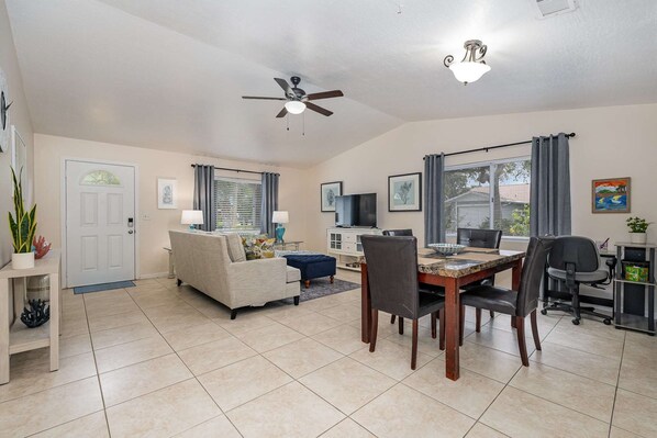 A spacious living room with a sofa, dining table, and chairs, a ceiling fan, and a TV.