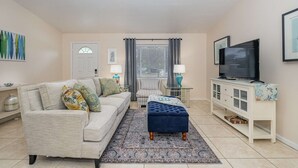 A living room with a sofa, ottoman, TV, and a cabinet. - Beachy Retreat 1 Mile to Beach (Satellite Beach)