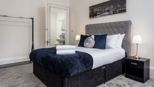A bedroom with a large bed, a nightstand, a wall-mounted lamp, and a cityscape picture. - St Meddans Mews in Troon (Troon)