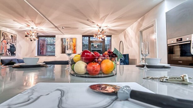 Une cuisine moderne avec un plan de travail en marbre, un panier de fruits et un coin repas agrémenté d’une nappe blanche.