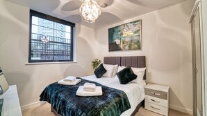 Une chambre à coucher avec un lit, une table de chevet, une fenêtre donnant sur des immeubles, un tableau au mur et un lustre.