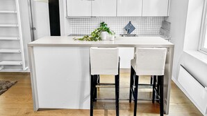 Una cocina moderna con armarios blancos, una isla central con dos taburetes altos, iluminación colgante y una planta en maceta.