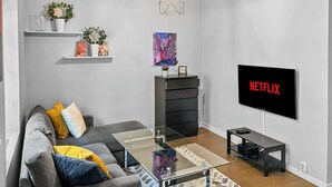 Una sala de estar moderna con un sofá gris, una mesa de centro de vidrio, un mueble de TV negro y un televisor montado mostrando 'NETFLIX'.