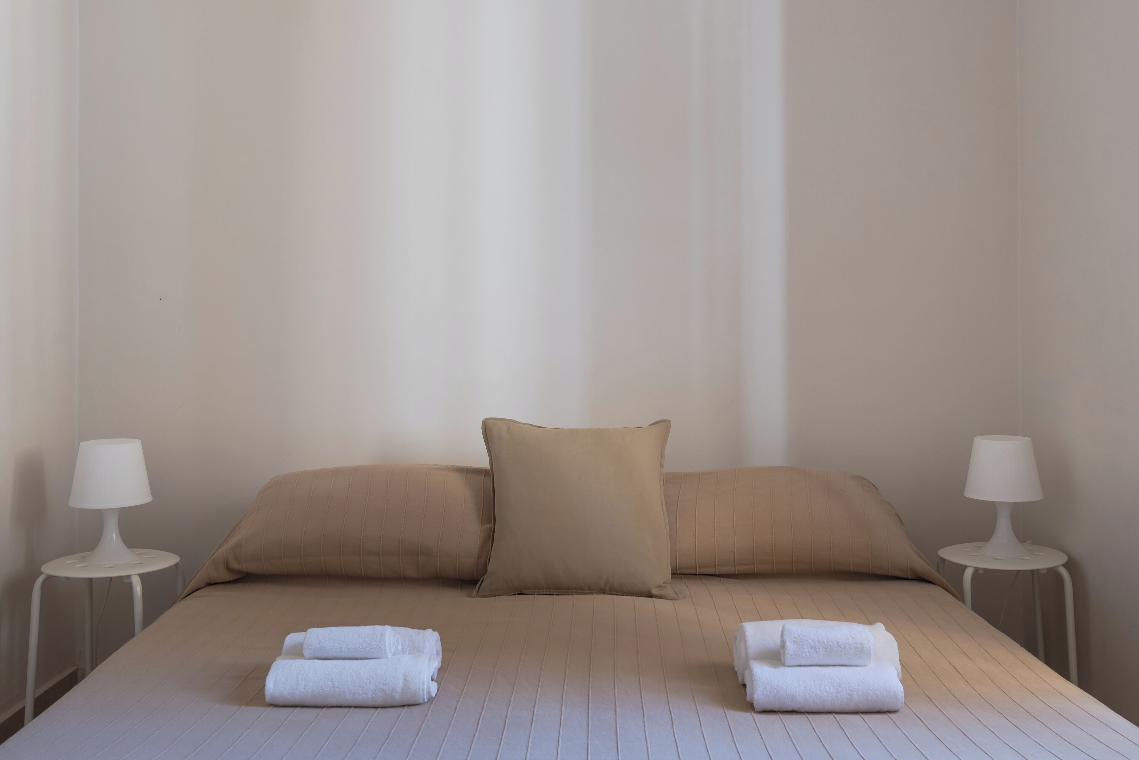 A neatly made bed with beige bedding and two pillows, flanked by two white bedside tables with lamps, in a minimalist room.