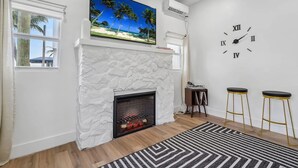A living room with a white stone fireplace, a large beach-themed painting above it, a wall clock, and two bar stools.