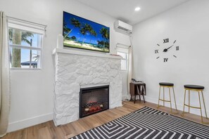 A living room with a white stone fireplace, a large beach-themed painting above it, a wall clock, and two bar stools.