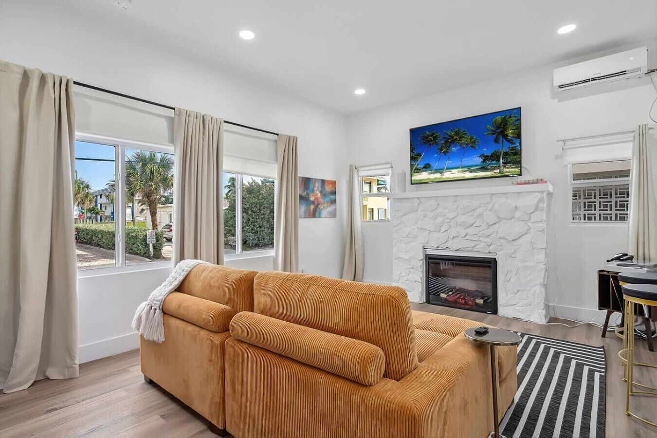 A living room with a sofa, a fireplace, a TV mounted on the wall, and a view of the outdoors through large windows.