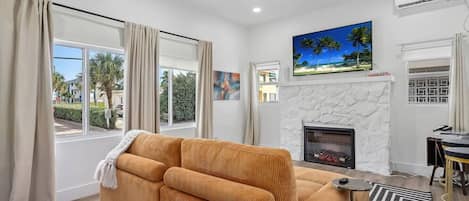 A living room with a sofa, a fireplace, a TV mounted on the wall, and a view of the outdoors through large windows.