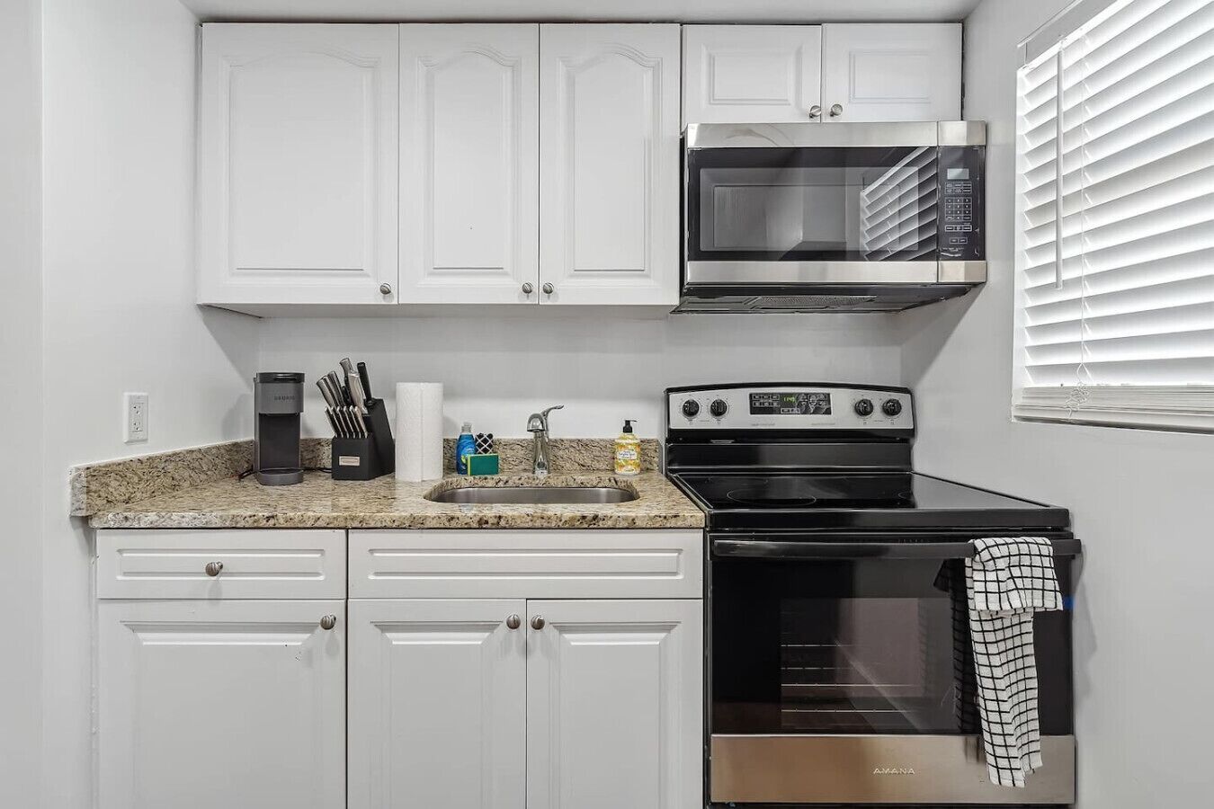 Una cocina con gabinetes blancos, un microondas de acero inoxidable, una estufa negra y un fregadero con varios objetos sobre la encimera.