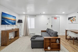Una sala moderna con un sofá gris, una mesa de centro de madera, un televisor en la pared y una puerta señalizada con