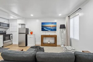 A modern living room with a grey sofa, a wooden coffee table, a flat-screen TV mounted above a wooden console, a white lamp, and a kitchen area with a fridge, oven, and microwave. - Casa Roselle 3 Bedrooms Beach access (HOLLYWOOD)