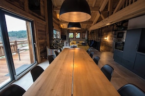 Een houten interieur met een lange eettafel, zwarte stoelen, een bank en een keukenhoek met moderne apparatuur.