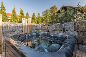 Un jacuzzi en un espacio exterior con paredes de piedra, una terraza de madera y una casa al fondo.