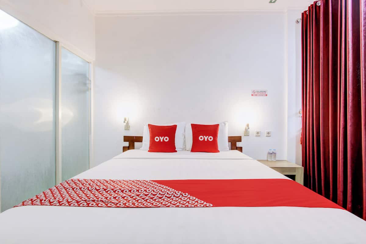 Ett hotellrum med en säng, två röda kuddar med ordet  ”OYO” på, ett rött och vitt mönstrat sängöverkast, ett träbord vid sängen med en flaska på och en röd gardin.