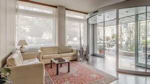 A spacious living room with a beige sofa, a small coffee table, and a red patterned rug. Large windows provide a view of the street outside. - Hotiday Celle Ligure (Celle Ligure)