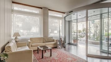 Un vaste espace de vie comprenant un canapé beige, une table basse et un tapis à motifs rouges. De grandes fenêtres offrent une vue sur la rue.
