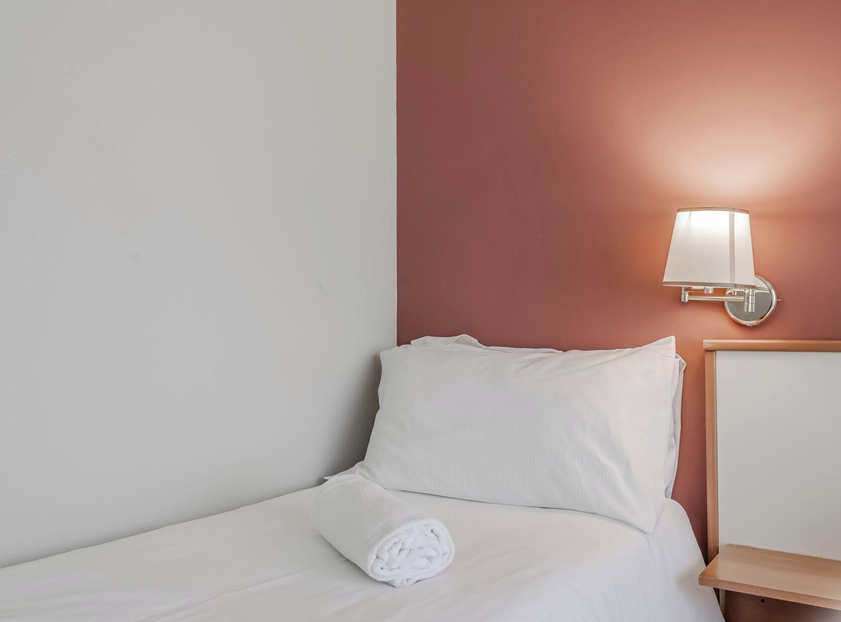 Een hotelkamer met een bed, een wandlamp en een netjes opgemaakt bed met een opgerolde handdoek.