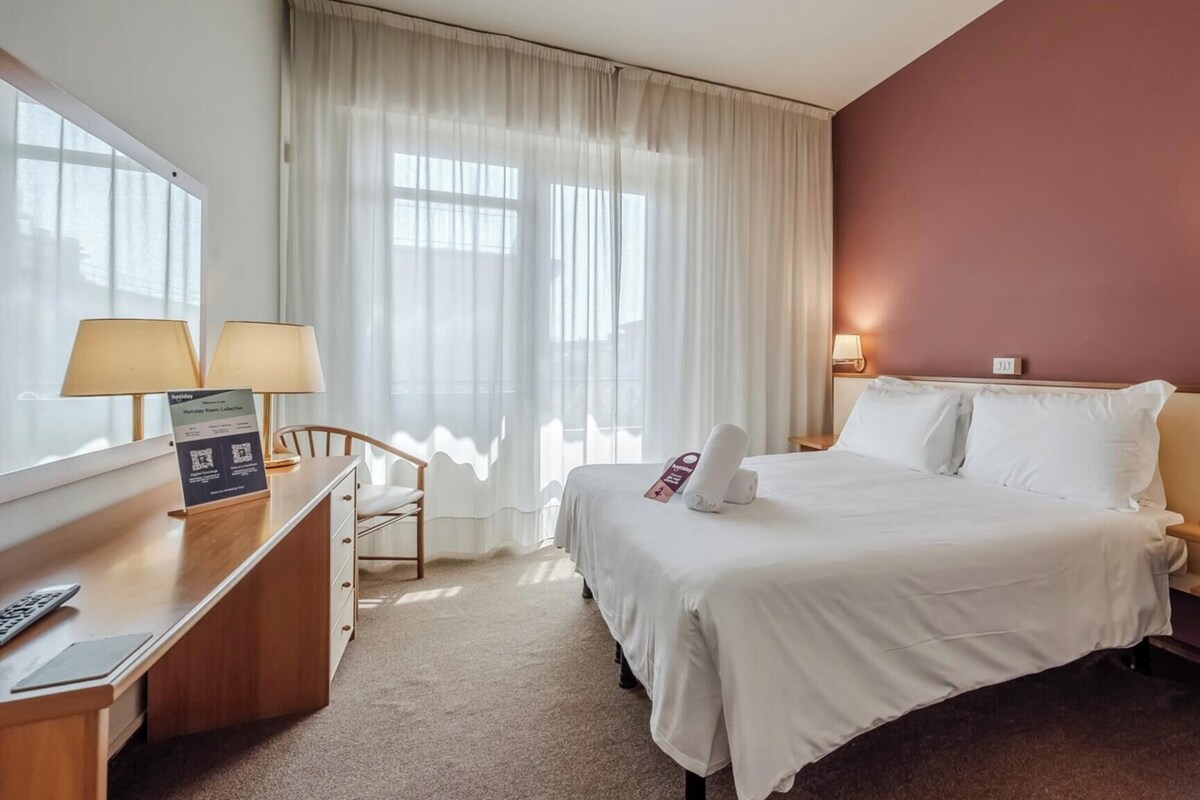 A hotel room with a bed, a desk with a chair, a lamp, and a window with curtains.