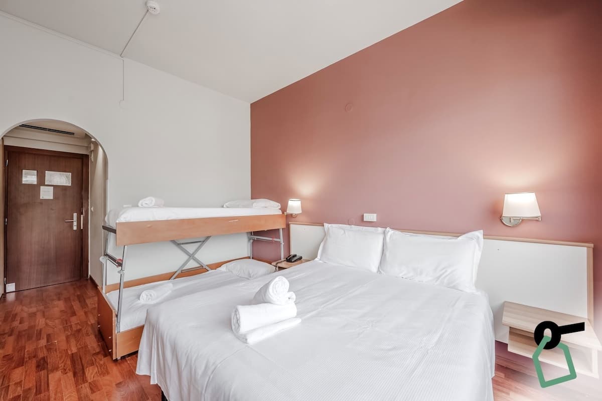 A hotel room with a bunk bed, a single bed, wooden flooring, a door with a notice, and two wall-mounted lamps.