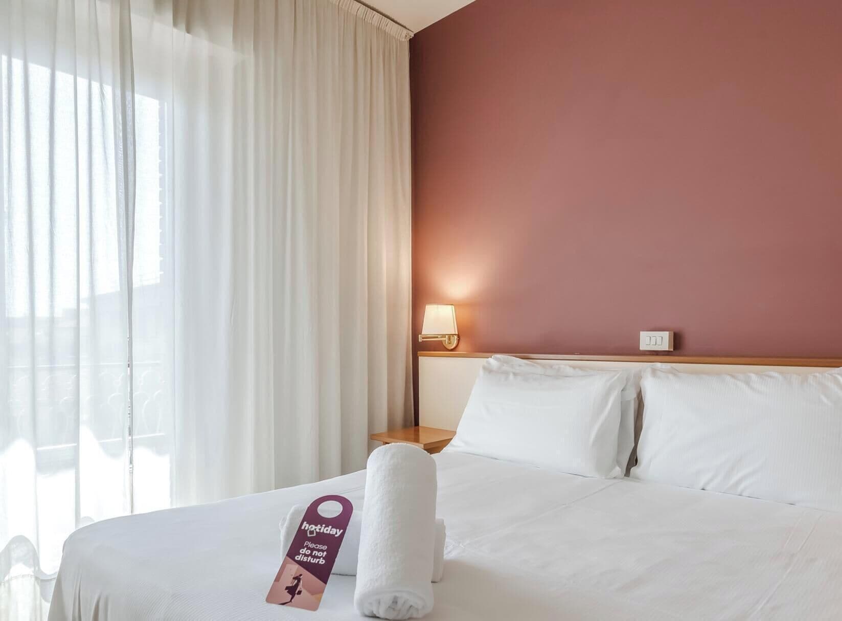 Una camera d'albergo con un letto, tende bianche, una lampada a muro e un cartello sul letto.