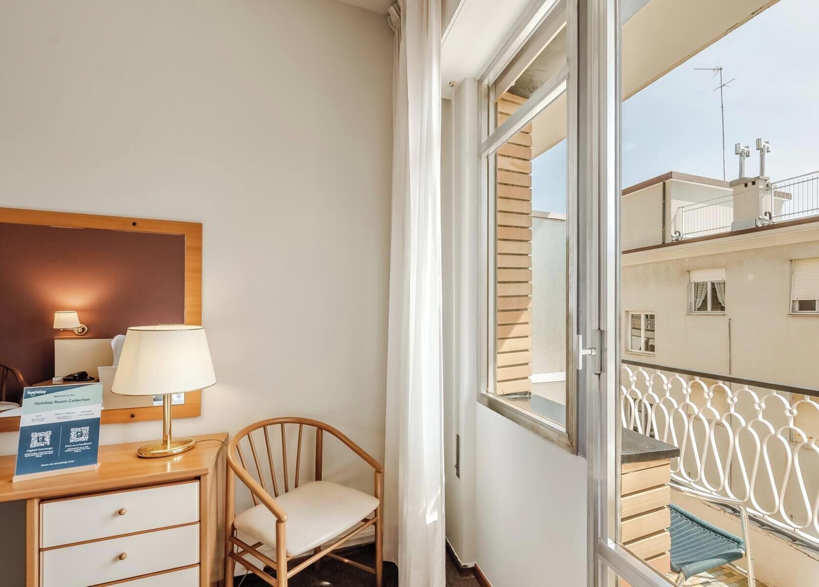 Une chambre d’hôtel équipée d’un bureau, d’une chaise, d’une lampe et offrant une vue sur la façade de l’immeuble.