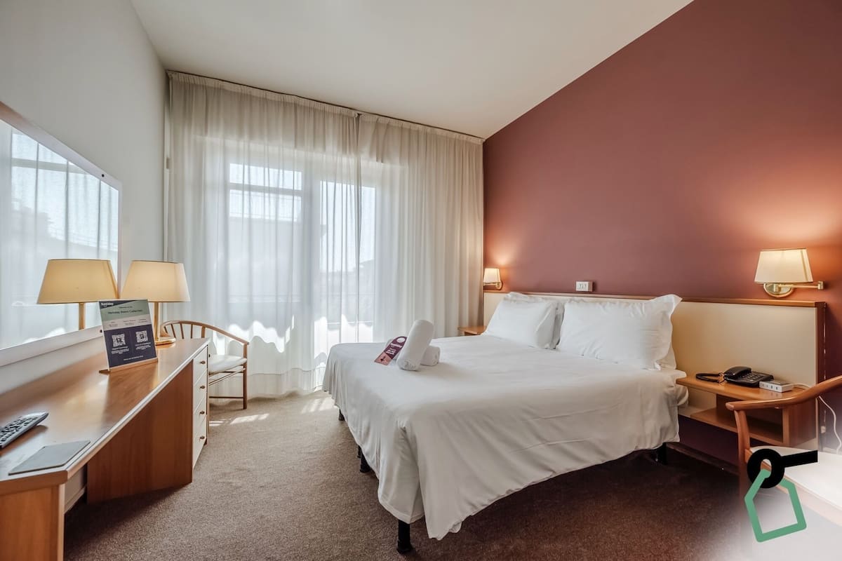 A hotel room with a bed, two bedside tables, a desk with a lamp, a chair, and a window with curtains.