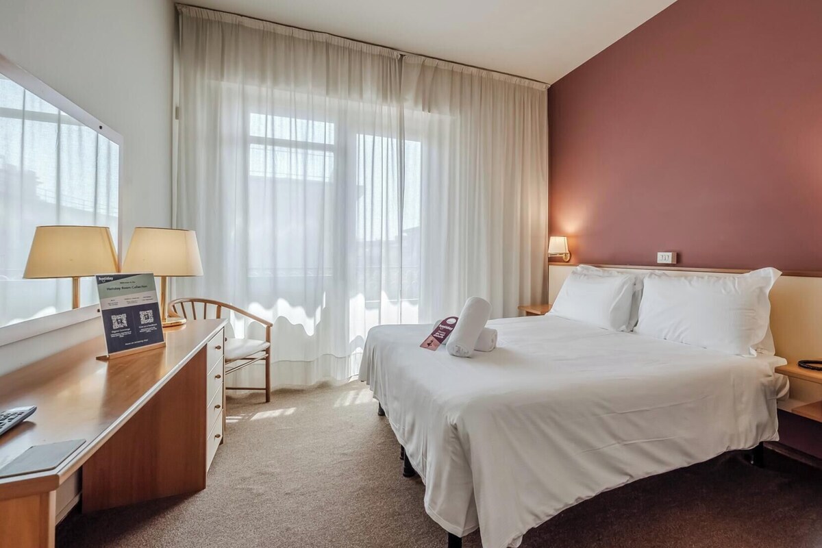 Habitación de hotel con cama, dos mesitas de noche, un escritorio con lámpara, una silla y una ventana con cortinas.