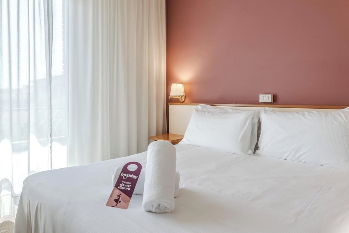 A hotel room with a bed, a nightstand, a lamp, and a window with curtains.